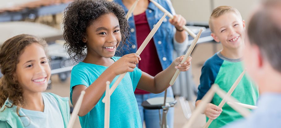 An unrecognizable band teacher stands in front of an attentive group of elementary age children and teaches drumming. The smiling children look at him and each hold a pair of drumsticks up in the air.