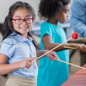 An adorable elementary age girl turns to smile for the camera as she stands beside her unrecognizable teacher and another little girl at a xylophone. She is learning to play the instrument in band class at school.